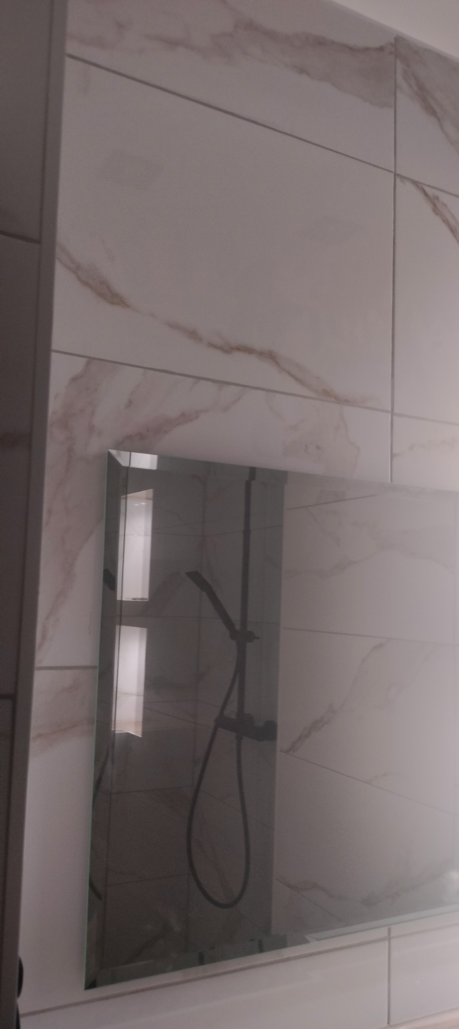 Wykończona ściana łazienki z białych płytek imitujących marmur, z lustrem odbijającym kabinę prysznicową z czarną armaturą.