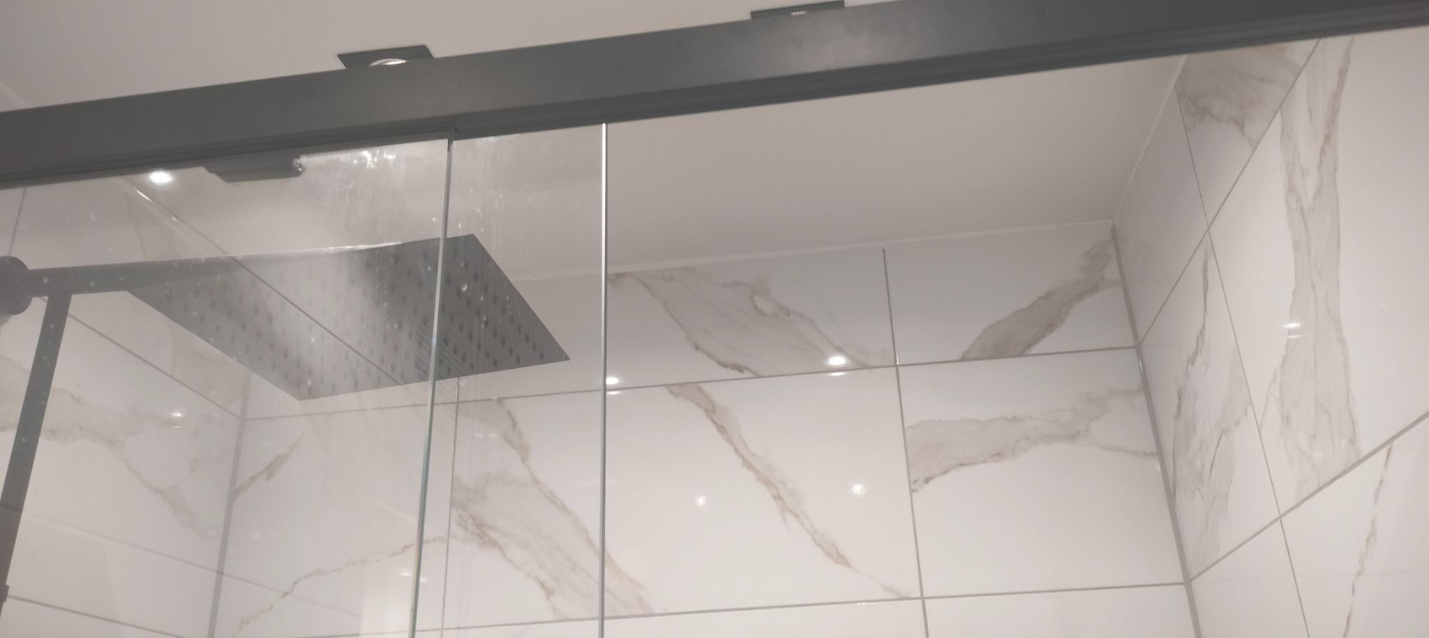 Kabina prysznicowa z deszczownicą, wykończona białymi płytkami imitującymi marmur, z szarymi profilami.