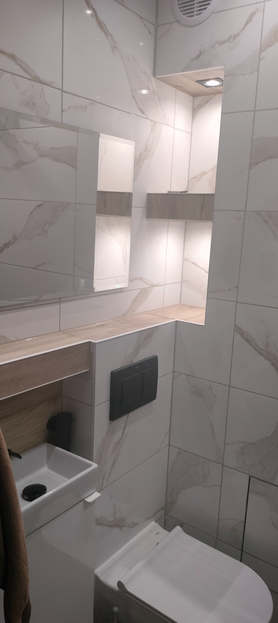 Nowoczesna łazienka z białymi płytkami imitującymi marmur, podświetlane wnęki, umywalka nablatowa i wisząca toaleta z czarnym przyciskiem spłukującym.