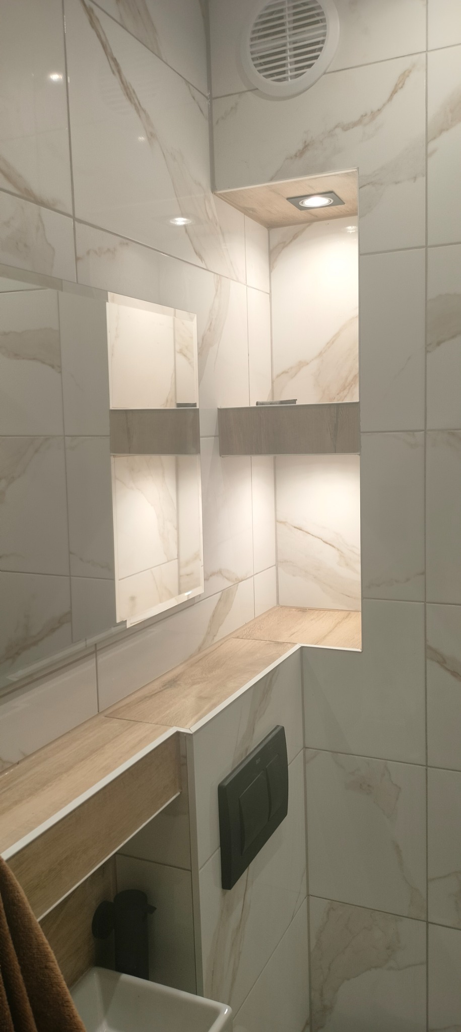 Nowoczesna łazienka wykończona białymi płytkami imitującymi marmur, z drewnianymi akcentami w postaci półek wnękowych podświetlonych punktowym światłem i blatu nad umywalką, widoczny czarny...