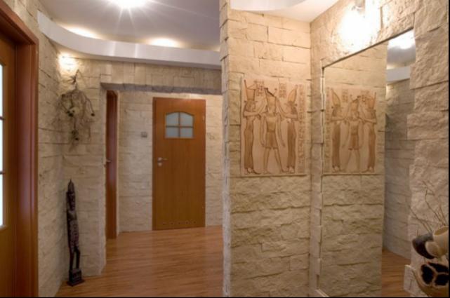 Korytarz wykończony kamieniem dekoracyjnym w odcieniach beżu, z drewnianą podłogą i drzwiami, ozdobiony egipskimi płaskorzeźbami i nowoczesnym oświetleniem.