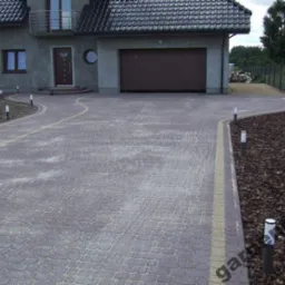 Ułożona kostka brukowa w odcieniach szarości i czerwieni tworząca podjazd do domu z garażem. Wzdłuż ścieżki rozmieszczone małe lampki oświetleniowe. Krawędzie wykończone jaśniejszą kostką. Obok...