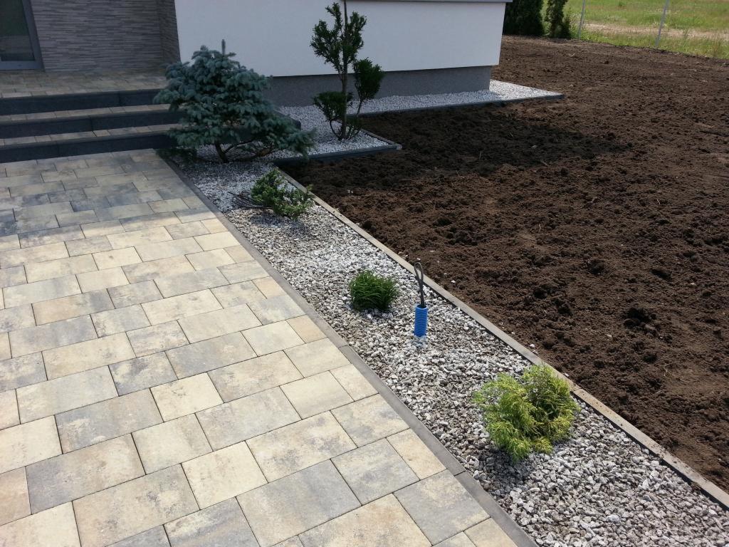 Ułożona kostka brukowa prowadząca do schodów wejściowych domu z rabatą z kamyków, roślinami i przygotowanym podłożem pod trawnik.