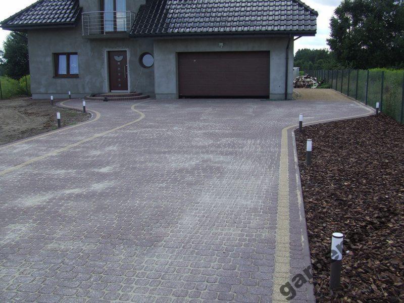 Ułożona kostka brukowa w odcieniach szarości i czerwieni tworząca podjazd do domu z garażem. Wzdłuż ścieżki rozmieszczone małe lampki oświetleniowe. Krawędzie wykończone jaśniejszą kostką. Obok...