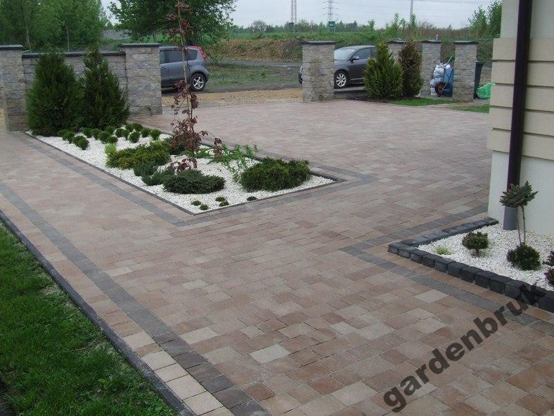 Ułożona kostka brukowa w odcieniach brązu i szarości, z dekoracyjnymi obrzeżami, rabatami z białym grysem i zielenią, oraz kamiennymi słupkami bramy w tle.