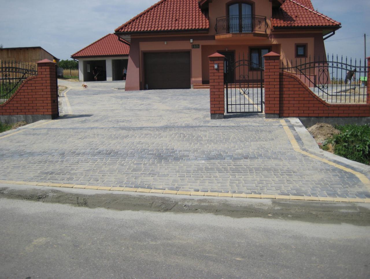 Ułożona szara kostka brukowa z żółtymi obrzeżami prowadząca do domu z czerwoną dachówką i kutą bramą wjazdową.