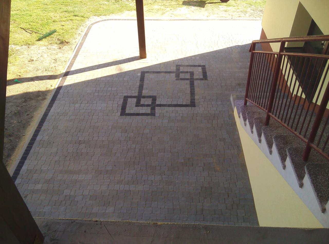 Ułożona kostka brukowa w odcieniach szarości z dekoracyjnym wzorem z ciemniejszych kostek, widok z góry, częściowo zacieniona przez konstrukcję budynku, obok schody z metalową barierką.