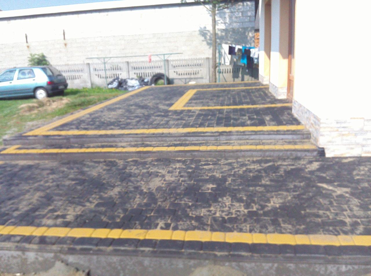 Ułożona kostka brukowa w kolorze czarnym z żółtymi obramowaniami na schodach i chodniku, widok z poziomu gruntu, w tle zaparkowany samochód i pranie na sznurze.