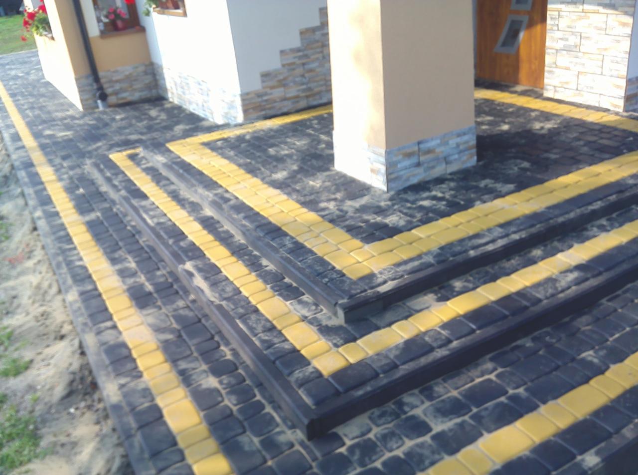 Ułożona kostka brukowa w kolorze grafitowym z żółtymi akcentami tworząca schody wejściowe do budynku, widoczne detale wykończenia elewacji.
