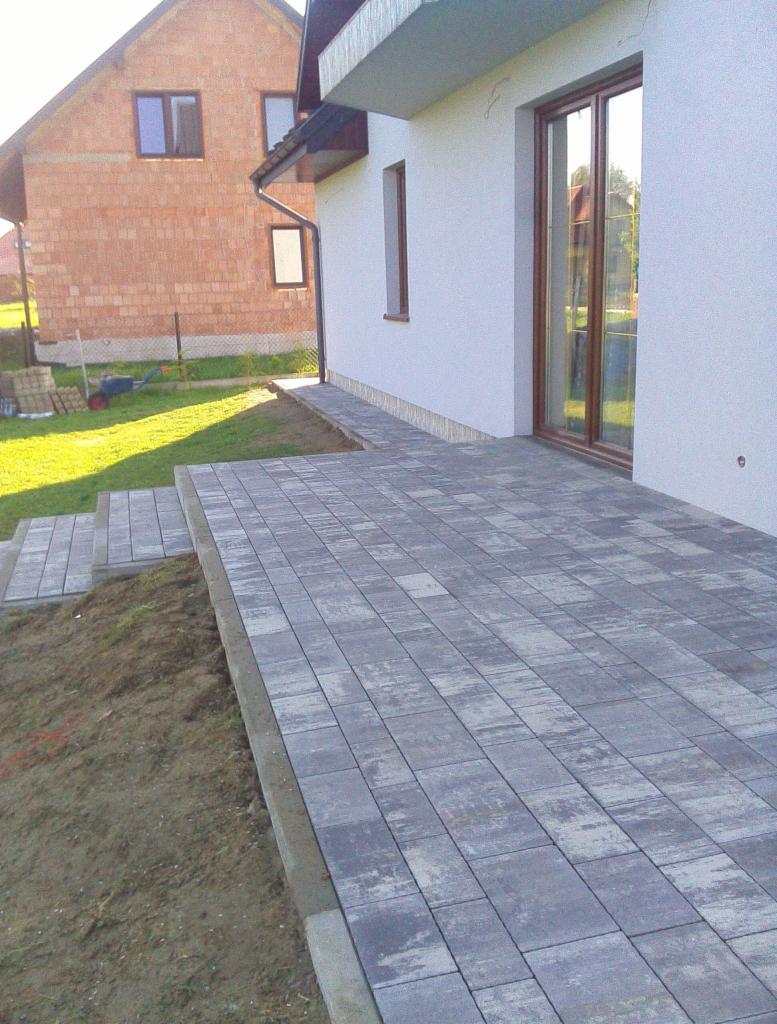 Szara kostka brukowa ułożona przy białym domu z brązowymi oknami; widoczne schodki z kostki i fragment trawnika z budynkiem w tle.