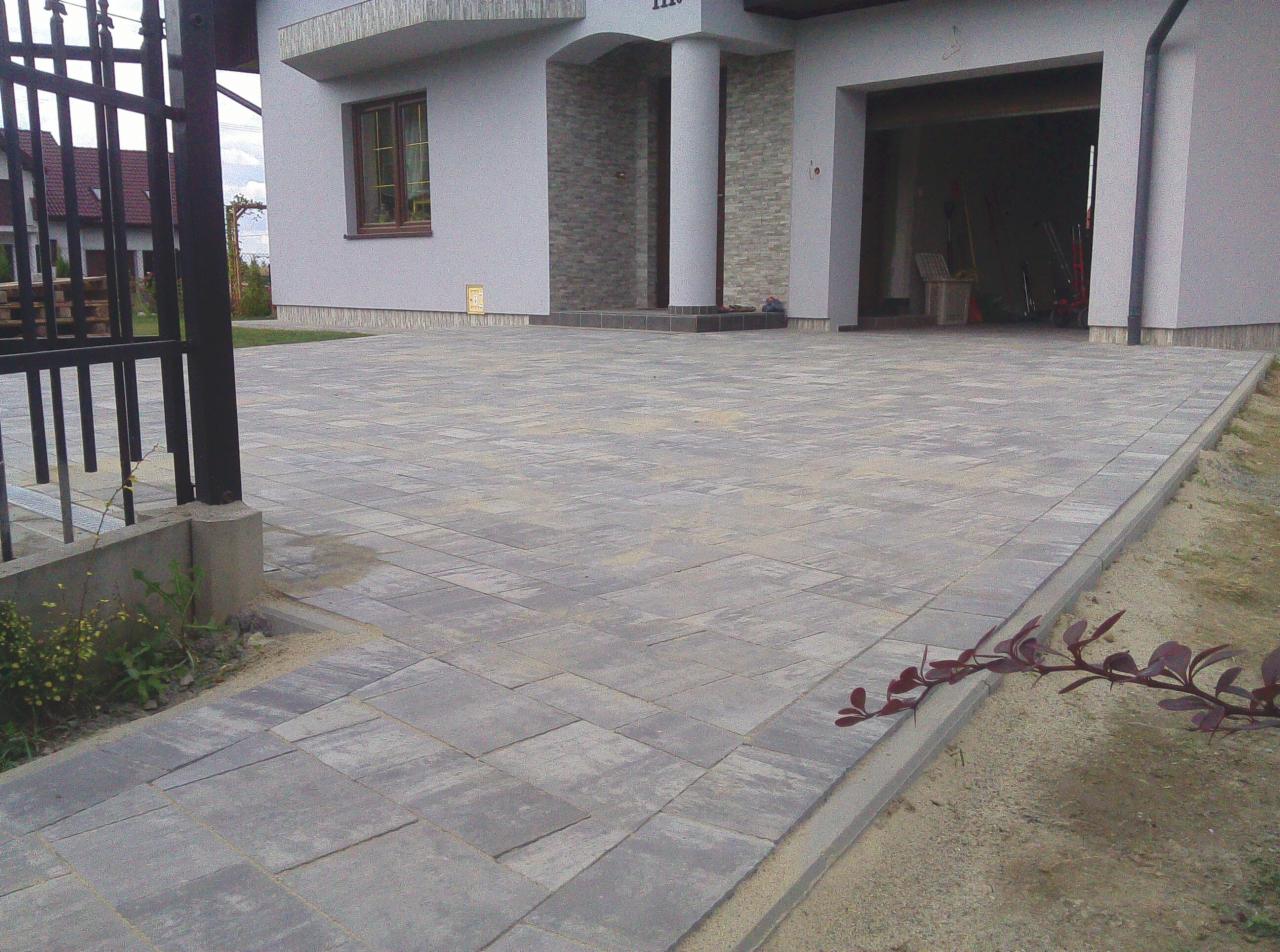 Szara kostka brukowa ułożona na podjeździe do domu jednorodzinnego, widok z poziomu gruntu na bramę wjazdową i otwarty garaż.