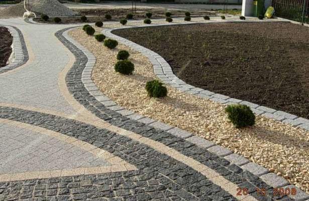 Kręta ścieżka z kostki brukowej w odcieniach szarości i beżu, obrzeża z jasnych kostek, wysypana drobnym żwirem z nasadzeniami z małych krzewów, w tle przygotowany teren pod trawnik.