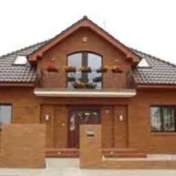 Elewacja domu jednorodzinnego z brązowej cegły, z balkonem zdobionym kwiatami i brązowym dachem z dachówki.