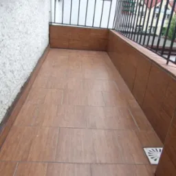 Wykończony balkon z brązowymi płytkami imitującymi drewno na podłodze i ścianach, widoczny odpływ wody. Balustrada z czarnymi metalowymi prętami.