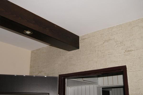 Fragment wnętrza: dekoracyjna ściana z deseniem imitującym pismo, ciemna belka stropowa z oświetleniem punktowym i rama drzwi w kolorze ciemnego brązu.