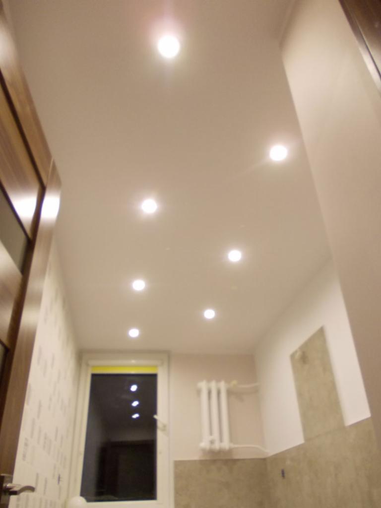 Biały sufit podwieszany z równomiernie rozmieszczonymi punktowymi lampami LED w wyremontowanym pomieszczeniu z oknem i grzejnikiem. Widoczny fragment drzwi z drewnianą ramą.