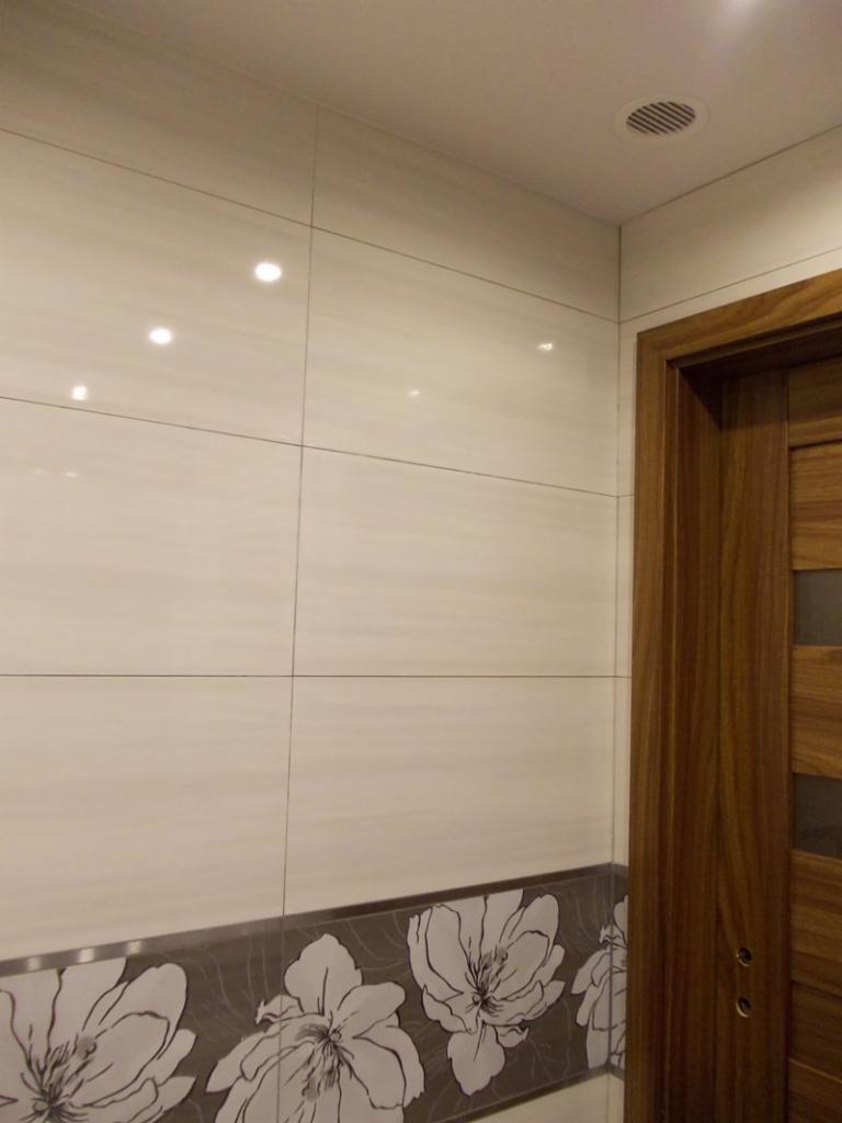 Wykończona łazienka: kremowe płytki na ścianach z motywem kwiatowym u dołu, drewniane drzwi po prawej stronie. Widoczny fragment sufitu z kratką wentylacyjną.