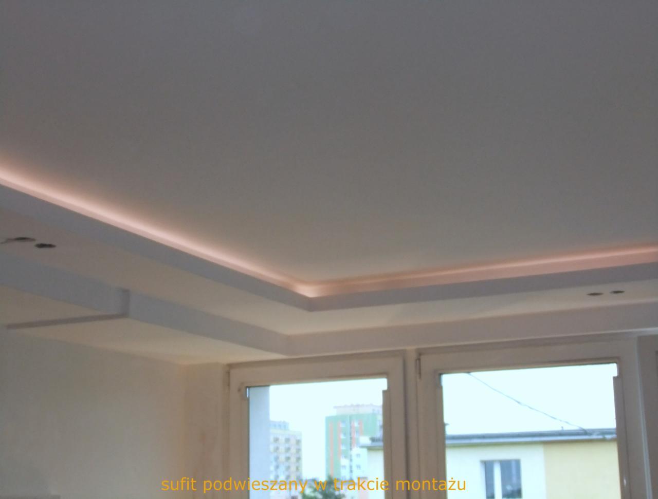 Biały sufit podwieszany z oświetleniem LED w trakcie montażu, widok z dołu, okno z widokiem na budynki w tle.