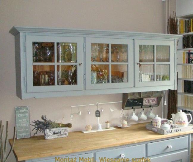 Jasnoszara szafka wisząca z przeszklonymi drzwiczkami nad blatem kuchennym. Widoczne naczynia wewnątrz, dekoracje na blacie i fragment regału z książkami.