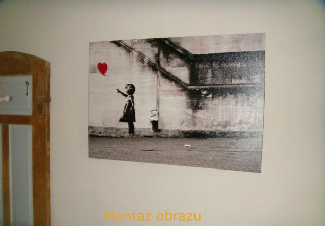 Zawieszony na ścianie obraz Banksy'ego z dziewczynką i czerwonym balonem w kształcie serca. Widoczny fragment ściany i drewniana rama drzwi.