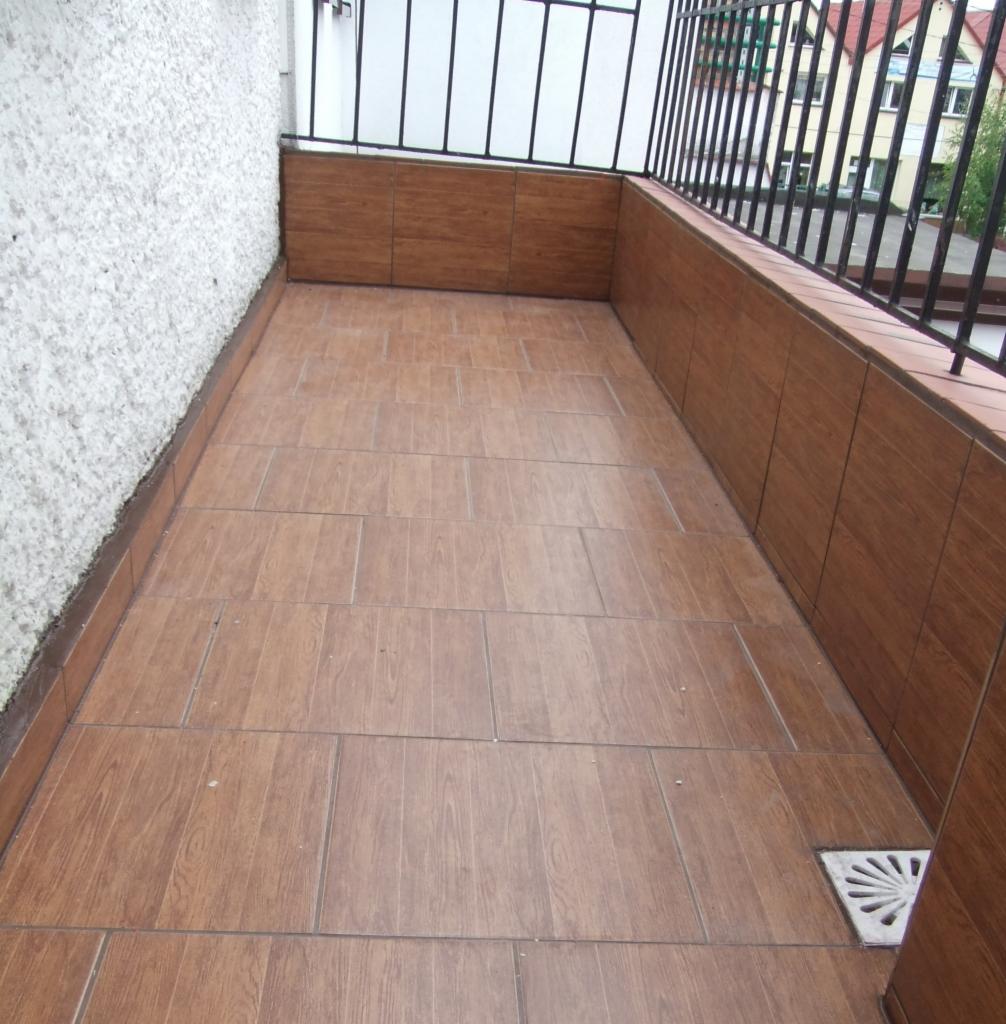Wykończony balkon z brązowymi płytkami imitującymi drewno na podłodze i ścianach, widoczny odpływ wody. Balustrada z czarnymi metalowymi prętami.