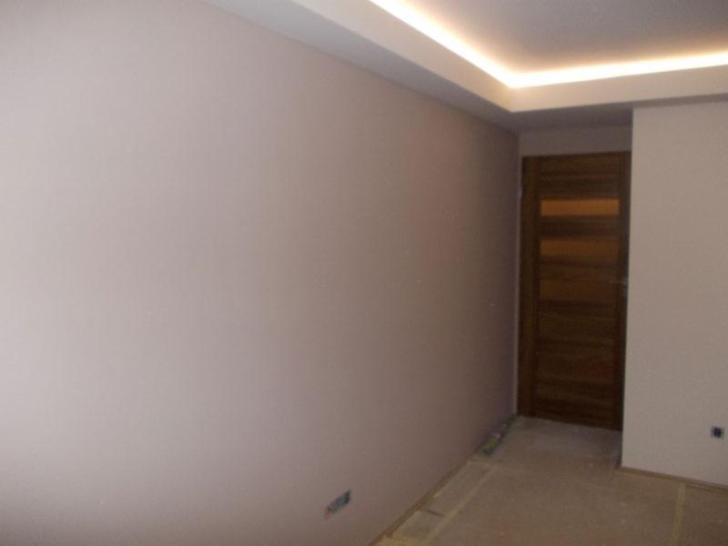 Pomieszczenie po malowaniu ścian na jasny kolor z drewnianymi drzwiami i oświetleniem sufitowym LED. Widoczne ślady po remoncie na podłodze.