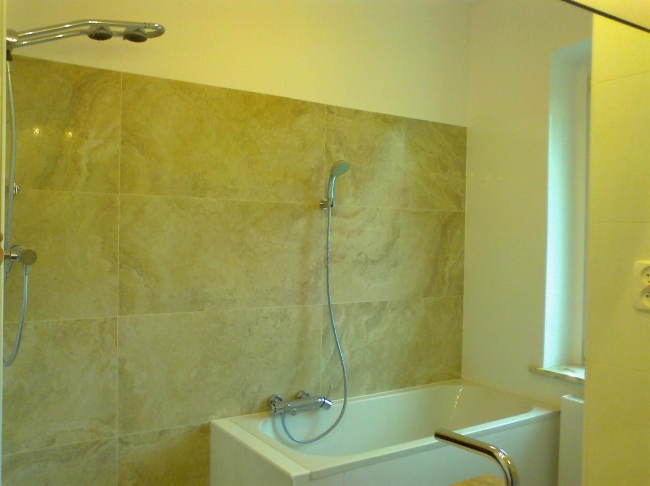 Wykończona łazienka z wanną, beżowymi płytkami imitującymi marmur i chromowaną armaturą. Widoczny prysznic deszczownica i standardowa słuchawka prysznicowa.