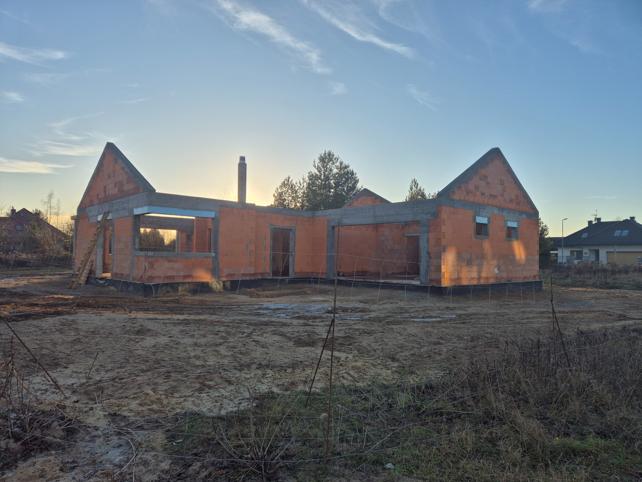 Wczesna faza budowy domu z czerwonej cegły, widoczne elementy konstrukcyjne, drabina oparta o ścianę, w tle drzewa i sąsiednie budynki, teren budowy ogrodzony siatką.