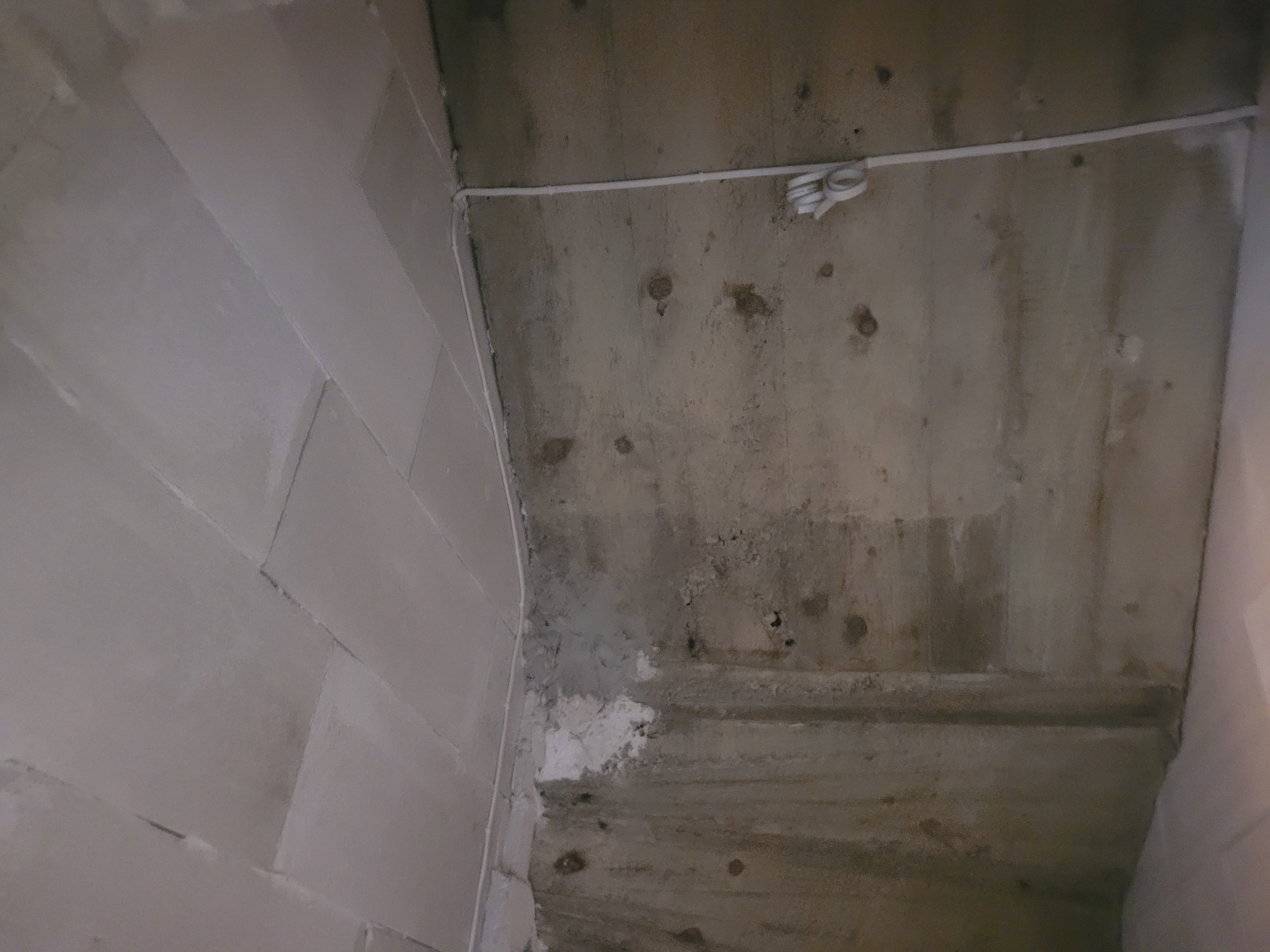 Surowy betonowy sufit z widocznymi śladami po szalunkach, częściowo pokryty białą gładzią i z poprowadzoną białą listwą elektryczną z luźno zwisającymi kablami.