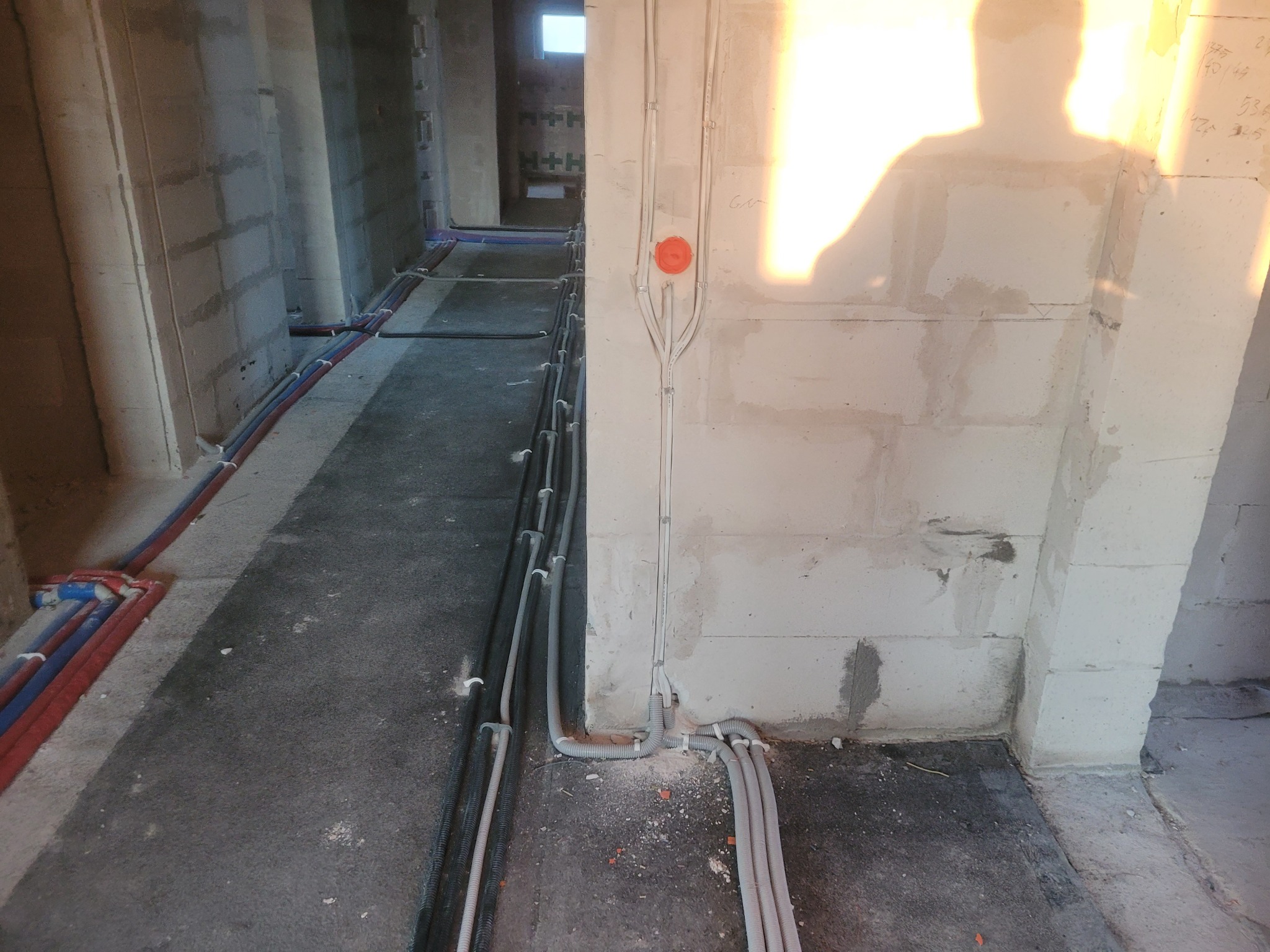 Wnętrze w trakcie budowy z widocznymi przewodami elektrycznymi ułożonymi wzdłuż ścian i podłogi, zakończonymi pomarańczową puszką instalacyjną na ścianie, oraz rurami instalacji wodnej biegnącymi...