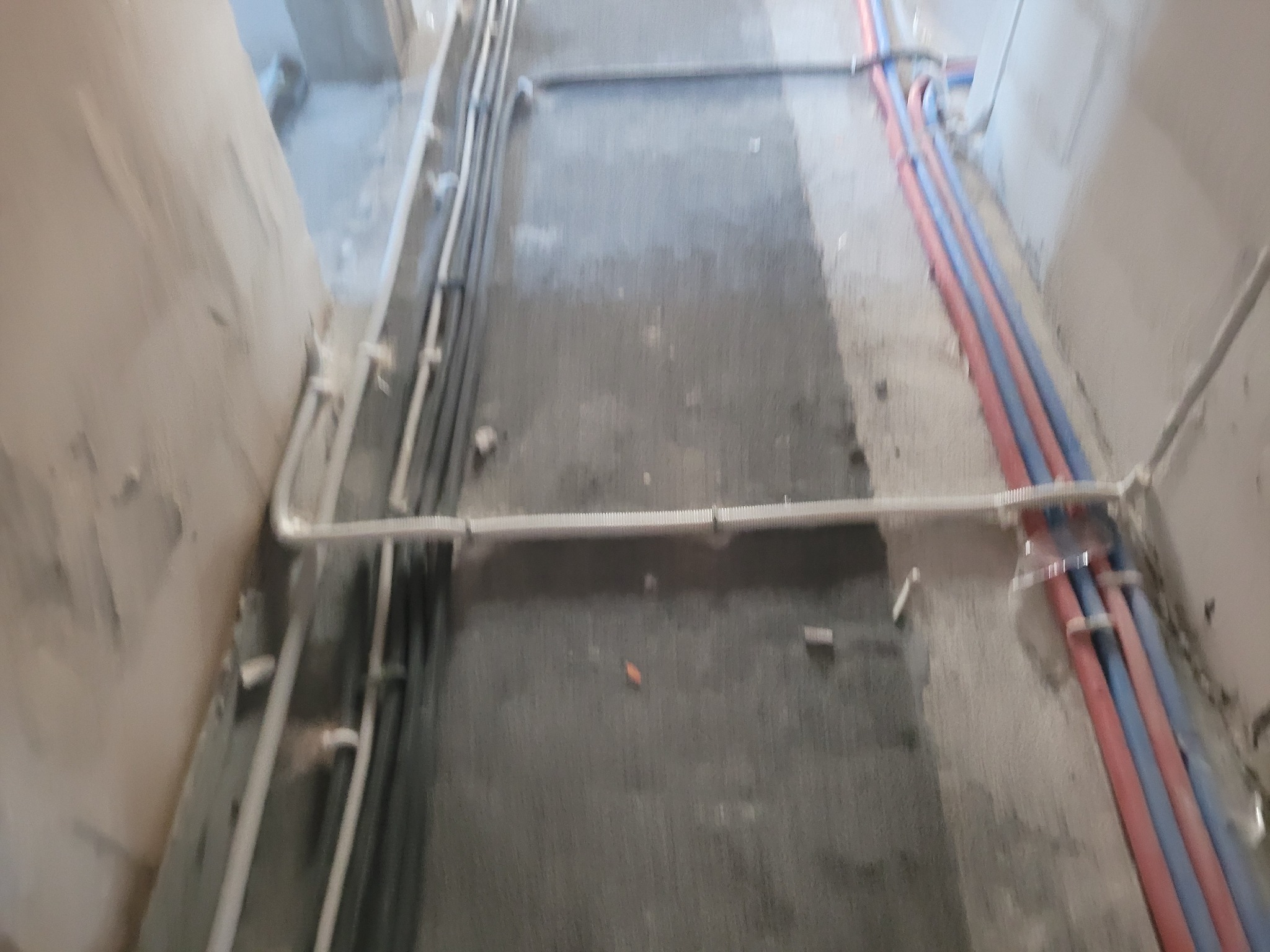 Wiązka rur w kolorze czerwonym i niebieskim, prowadzone wzdłuż ściany w surowym stanie, obok widoczne szare rury instalacji elektrycznej.