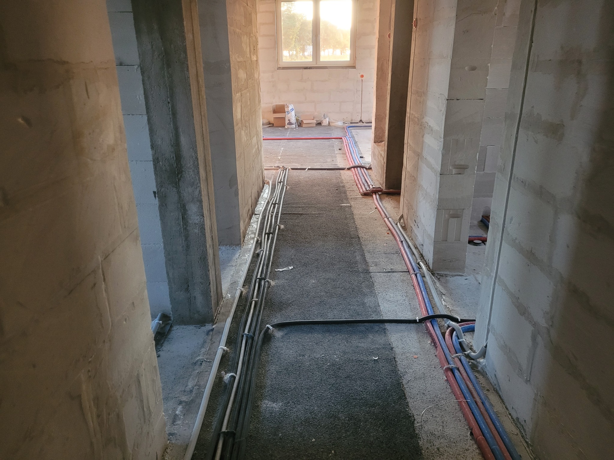 Wnętrze w stanie surowym z widocznymi instalacjami elektrycznymi i hydraulicznymi prowadzonymi po podłodze, z oknem w oddali wpadającym światłem słonecznym.