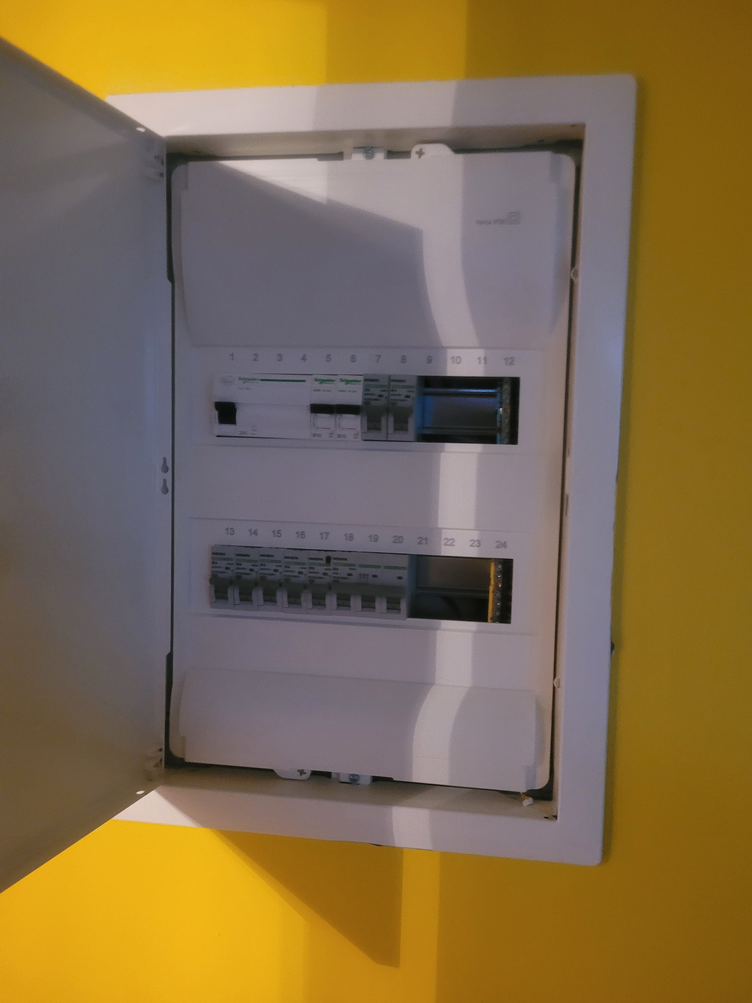 Otwarta skrzynka elektryczna z bezpiecznikami i wyłącznikami, zamontowana na żółtej ścianie, widoczne numery obwodów od 1 do 24.