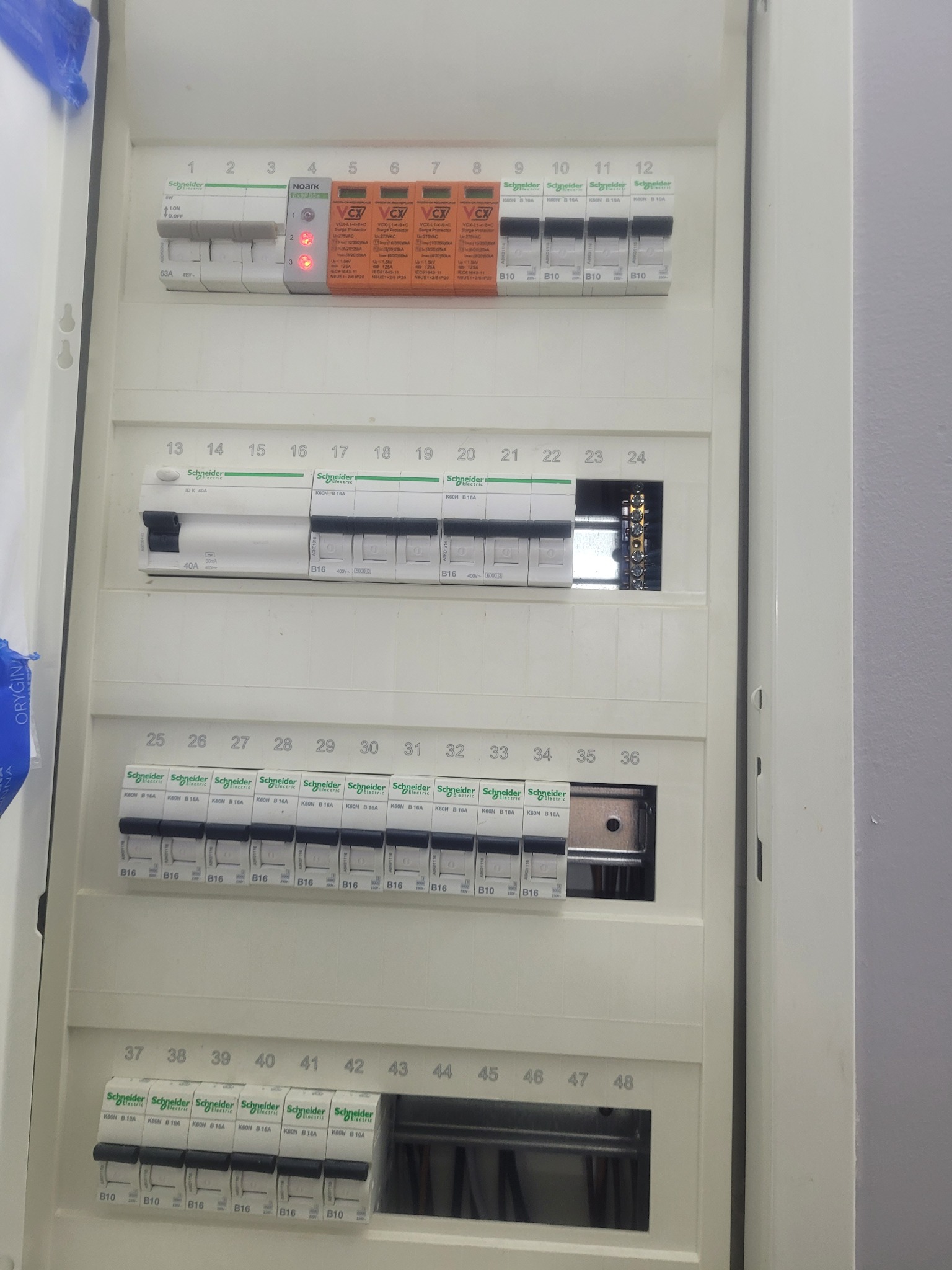 Otwarta skrzynka elektryczna z widocznymi zabezpieczeniami przepięciowymi, wyłącznikami różnicowoprądowymi i nadprądowymi marki Schneider Electric i Noark, oznaczona numerami od 1 do 48.