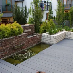 Nowoczesny ogród z tarasem z kompozytu, kaskadą wodną obłożoną kamieniem naturalnym i podwyższonymi rabatami z białej cegły, otoczony zielenią.