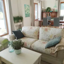 Jasny salon z sofą w kwiaty, drewnianym stołem kawowym i widokiem na jadalnię, akcenty w kolorze turkusowym.