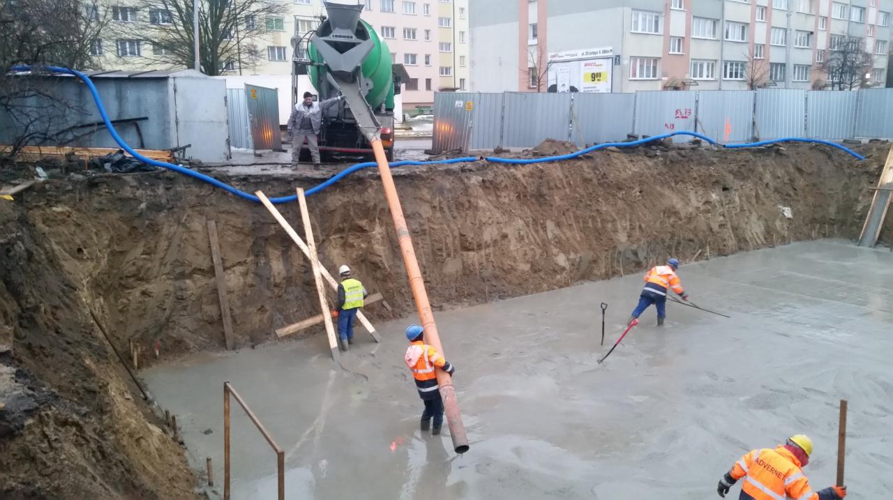 Wylewanie fundamentów pod budynek mieszkalny; betoniarka Liebherr podaje beton przez pomarańczową rurę, robotnicy w odblaskowych ubraniach rozprowadzają mieszankę, w tle ogrodzenie placu budowy...
