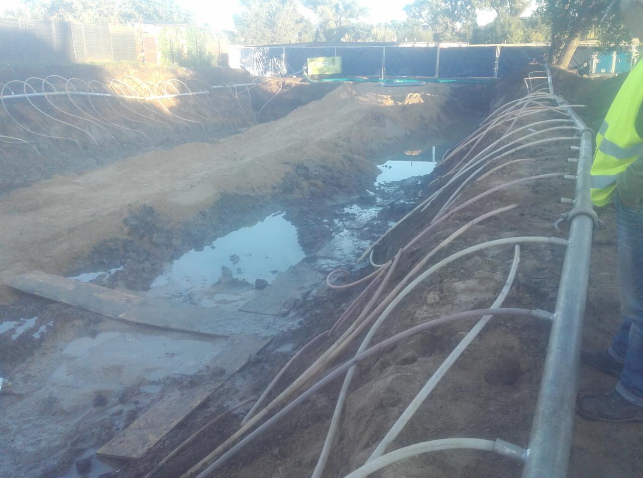 Wykop z wodą gruntową i ułożonymi przewodami rurowymi wzdłuż ścian, widoczny fragment pracownika w żółtej kamizelce odblaskowej.