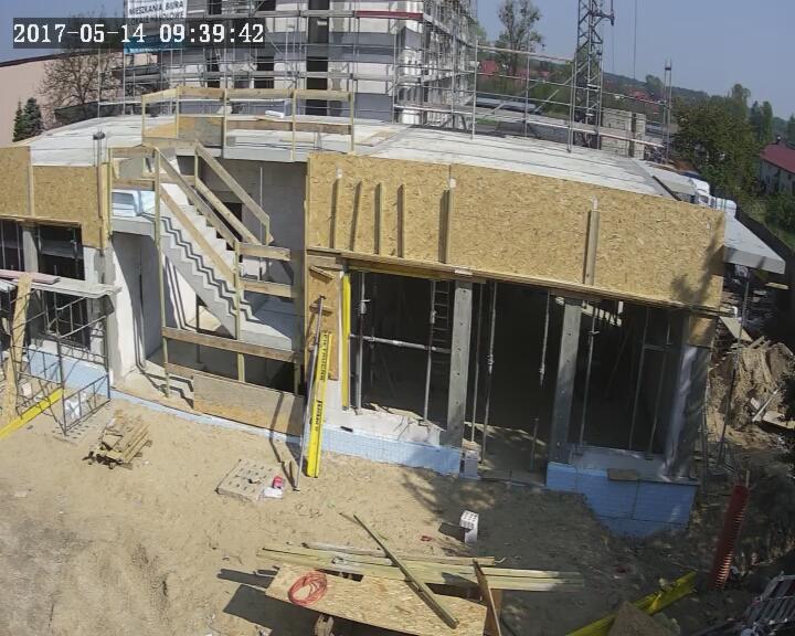 Budowa domu w trakcie realizacji, widoczne ściany z betonu komórkowego, drewniane szalunki, schody betonowe, rusztowania i materiały budowlane na placu budowy, ujęcie z góry.