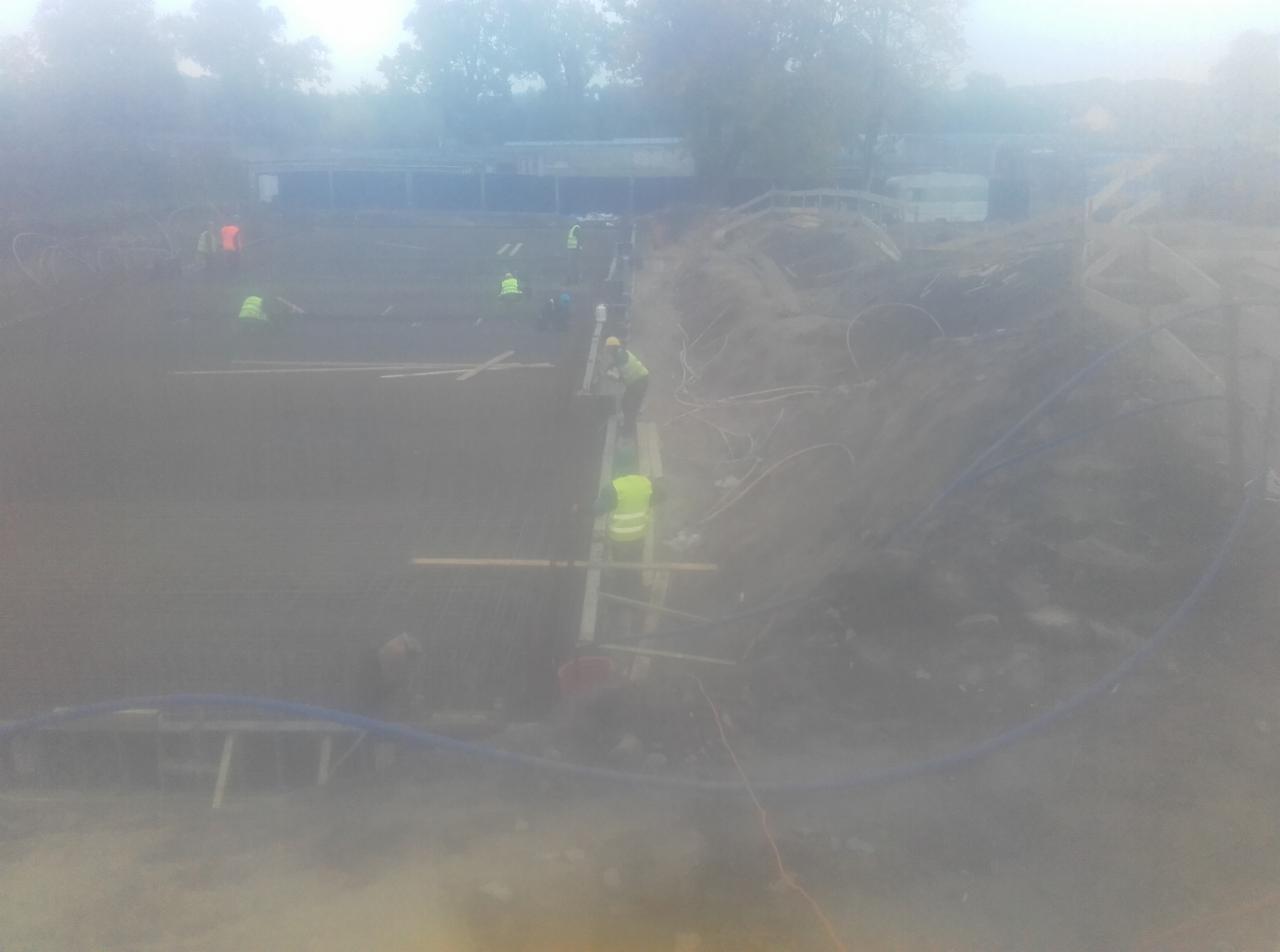 Budowa fundamentów domu: pracownicy w kamizelkach odblaskowych układają zbrojenie w wykopie, widoczne szalunki i niebieskie rury instalacyjne.