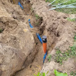 Wykop w ziemi z ułożonymi niebieskimi rurami osłonowymi i pomarańczową rurą kanalizacyjną z czarną pokrywą, widoczne wystające końcówki rur na trawie.