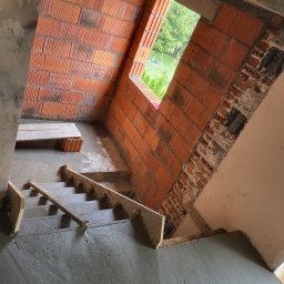 CONCRET JOB - Wnętrze w trakcie budowy: surowe betonowe schody z drewnianym szalunkiem, ściana z czerwonej cegły dziurawki z widocznym oknem na zewnątrz.