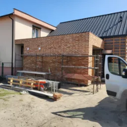 Budowa domu: fragment murowanej ściany z cegły na tle rusztowania, widoczny fragment elewacji z ociepleniem oraz samochód dostawczy