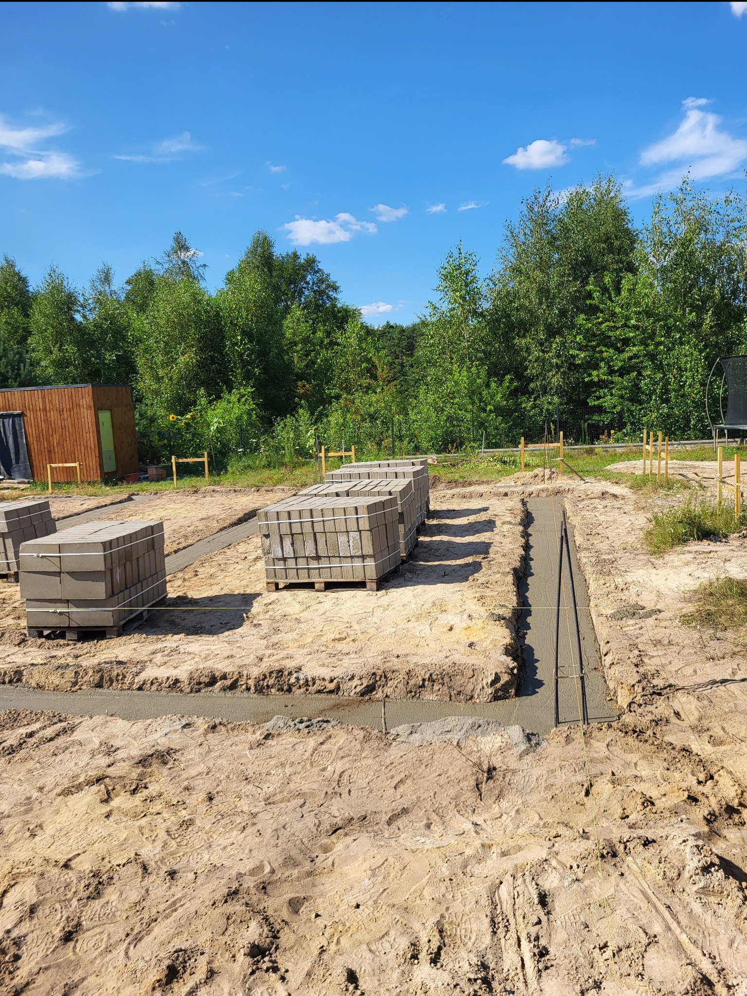 Przygotowany wykop z zalewanym betonem i ułożonym zbrojeniem pod fundament, obok palety z bloczkami betonowymi, teren budowy z drewnianym domkiem w tle.