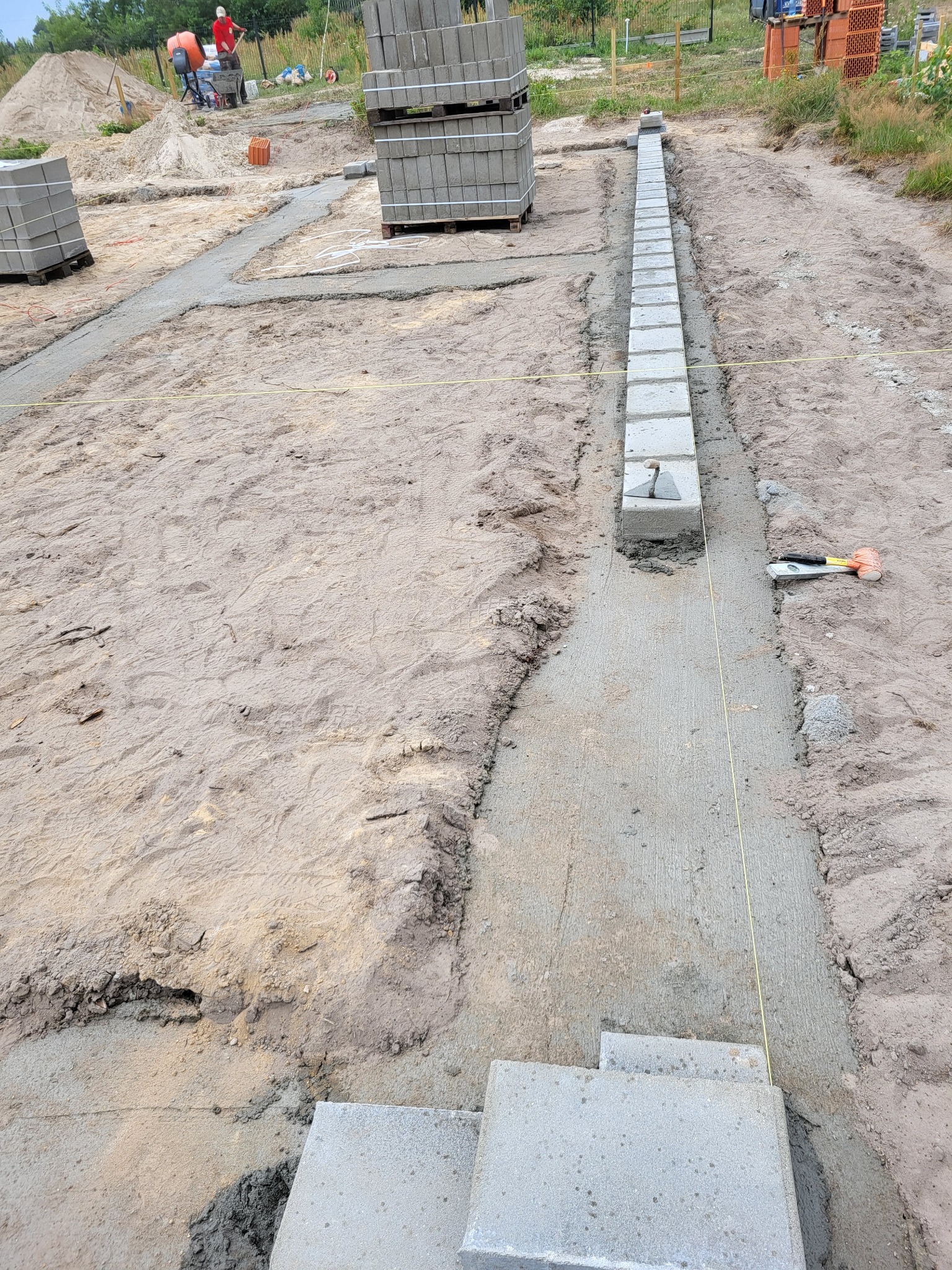 Budowa fundamentów: Wylany betonowy pas pod fundamenty z ułożonymi bloczkami betonowymi, widoczne narzędzia murarskie i wytyczona linia. W tle pracownicy budowlani i zapas bloczków.