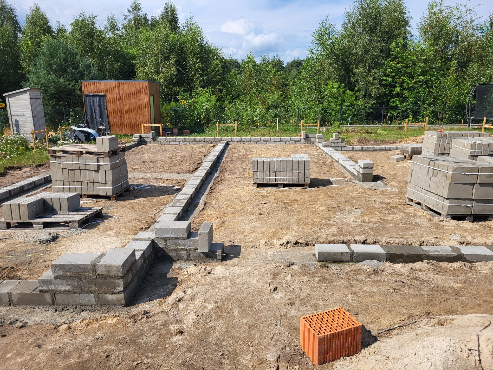 Wykop pod fundamenty z częściowo wymurowanymi ścianami z szarych bloczków betonowych, palety bloczków w tle, w oddali drewniana altana i drzewa.