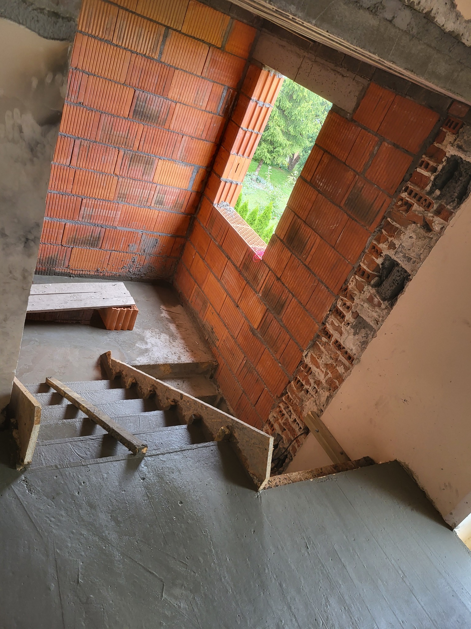 Wnętrze w trakcie budowy: surowe betonowe schody z drewnianym szalunkiem, ściana z czerwonej cegły dziurawki z widocznym oknem na zewnątrz.