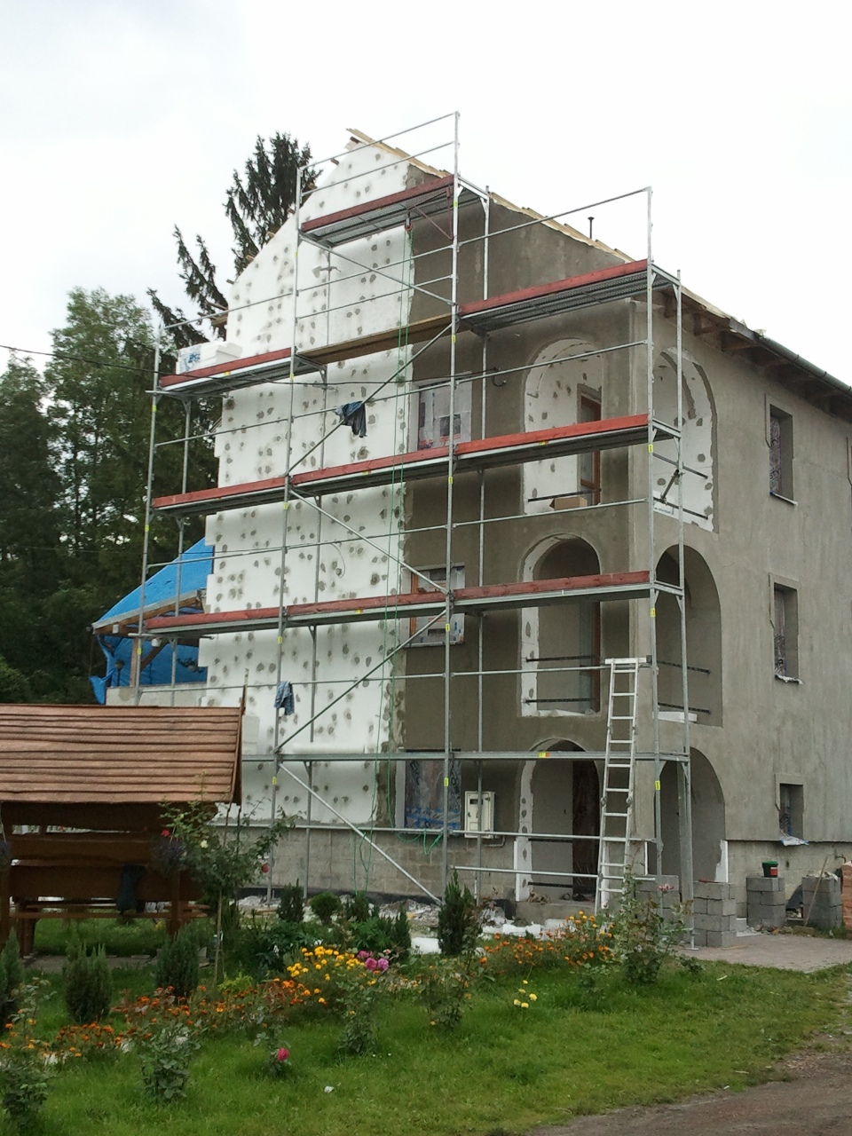 Budynek w trakcie docieplania elewacji styropianem, z widocznymi rusztowaniami i elementami ogrodu w tle.