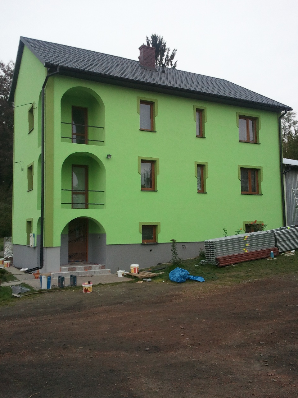 Dwukondygnacyjny dom z elewacją w odcieniach zieleni, z balkonami w stylu arkadowym, widoczne ślady farby i materiały budowlane wokół domu, sugerujące prace wykończeniowe.