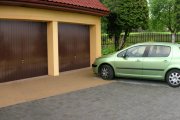 Dwu-stanowiskowy garaż z brązowymi, segmentowymi drzwiami garażowymi, pomalowany na beżowo, z zielonym samochodem osobowym zaparkowanym przed wjazdem.